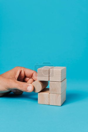 Foto de Un hombre pone un bloque de juguete cilíndrico en una pila de bloques de juguete rectangulares, sobre un fondo azul con un poco de espacio en blanco en la parte superior - Imagen libre de derechos