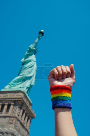 Foto de Un hombre levanta el puño, llevando una pulsera con una bandera de orgullo gay, frente a Lady Liberty, en Liberty Island, Nueva York, Estados Unidos - Imagen libre de derechos