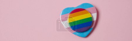 Foto de Una insignia redonda estampada con la bandera del arco iris colocada en una insignia en forma de corazón estampada con la bandera del orgullo transgénero, sobre un fondo rosa, en un formato panorámico para usar como banner web - Imagen libre de derechos