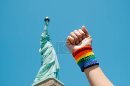 Photo for Man raising his fist, wearing a wristband patterned with a gay pride flag, in front of the Statue of Liberty, in Liberty Island, New York, United States - Royalty Free Image