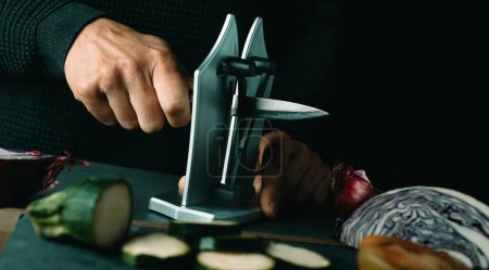 Photo for Man sharpening a kitchen knife with a desktop sharpener on a table, in a panoramic format to use as web banner or header - Royalty Free Image