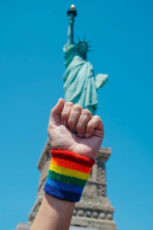 Téléchargez les photos : Gros plan d'un homme levant son poing, avec un bracelet orné d'un drapeau arc-en-ciel, devant la Statue de la Liberté, à New York, États-Unis - en image libre de droit