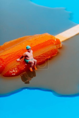 Téléchargez les photos : Gros plan d'un homme miniature, portant un maillot de bain vert, un bonnet de bain et une paire de lunettes de bain, assis sur une glace orange fondante pop sur un fond bleu - en image libre de droit