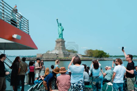 Foto de Ciudad de Nueva York, Estados Unidos - 22 de mayo de 2023: Un barco de turismo navegando frente a la Estatua de la Libertad, en Liberty Island, Nueva York, Estados Unidos, en un soleado día de primavera - Imagen libre de derechos
