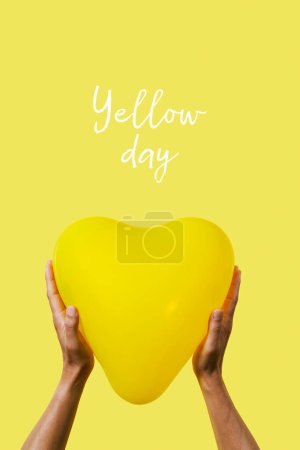 Photo for Closeup of a man holding a yellow heart-shaped balloon and text yellow day on a yellow background - Royalty Free Image