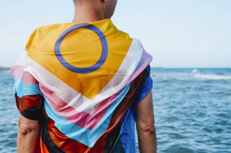 Téléchargez les photos : Une personne humide se tient dans la mer enveloppée dans un drapeau de fierté de progrès intergenre inclusif, vu de derrière - en image libre de droit