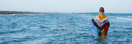 Téléchargez les photos : Une personne debout dans la mer enveloppée dans un drapeau de fierté de progrès intergenre humide, vu de derrière, dans un format panoramique à utiliser comme bannière Web - en image libre de droit