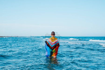 Téléchargez les photos : Une personne se tient dans la mer enveloppée dans un drapeau de fierté de progrès intersexuée humide debout dans l'eau, vu de derrière, un jour d'été - en image libre de droit