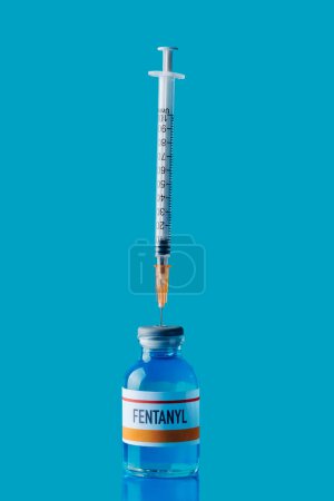a simulated vial of fentanyl with a syringe in its rubber stopper on a blue surface against a blue background