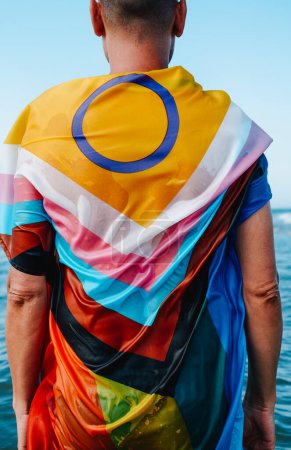 Photo for A wet person, seen from behind, wrapped in an intersex-inclusive progress pride flag standing in the seaa - Royalty Free Image