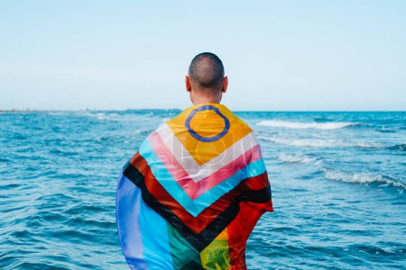 Foto de Un hombre mojado se para en el océano, mirando hacia el horizonte, envuelto en una bandera de orgullo de progreso intersex-inclusive mojado, visto por detrás - Imagen libre de derechos