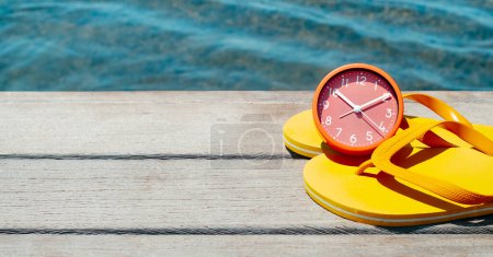 Téléchargez les photos : Une paire de tongs jaunes et une horloge rose sur une jetée en bois, à côté de l'eau, dans un format panoramique à utiliser comme bannière Web ou en-tête - en image libre de droit