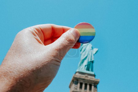 Photo for A man is holding a badge patterned with the rainbow flag in front of the Statue of Liberty, in New York, United States, on a sunny spring day - Royalty Free Image