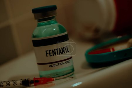 Téléchargez les photos : Closeup of a simulated vial of fentanyl and a syringe on a toilet cistern next to a cigarette butt, in a bathroom with dramatic and sordid lighting - en image libre de droit