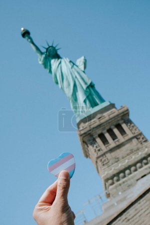 Téléchargez les photos : Une personne porte un insigne en forme de cœur orné du drapeau de la fierté transgenre, devant la Statue de la Liberté, alias Lady Liberty, à New York, États-Unis - en image libre de droit