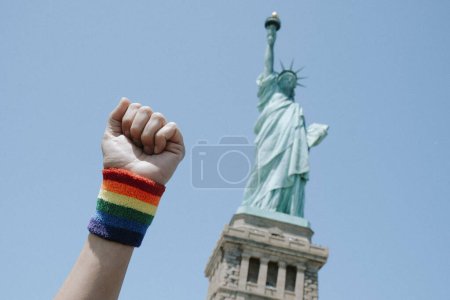 Téléchargez les photos : Un homme lève le poing, portant un bracelet orné du drapeau arc-en-ciel, devant la Statue de la Liberté, à New York, États-Unis - en image libre de droit