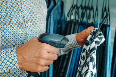 Foto de Un asistente de tienda en una tienda de ropa utiliza un escáner de código de barras portátil para escanear la información de una camisa que cuelga de una percha - Imagen libre de derechos