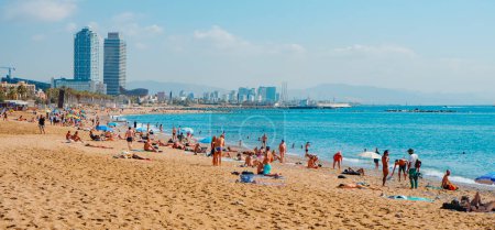Foto de Barcelona, España - 27 de julio de 2023: bañistas y bañistas en la playa de Sant Sebastia en Barcelona, España, temprano en la mañana de un día de verano, y las torres gemelas Hotel Arts y Torre Mapfre en el fondo - Imagen libre de derechos