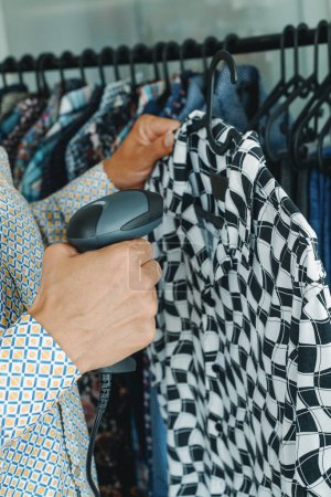 Photo for A shop assistant in a clothes shop is using a barcode scanner to scan the information of a shirt hanging from a hanger - Royalty Free Image