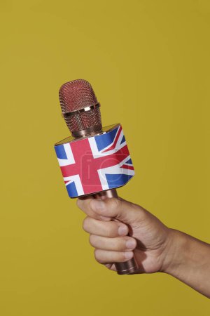 Téléchargez les photos : Gros plan de la main d'un homme tenant un microphone orné de la prise syndicale, drapeau du royaume uni, sur fond jaune - en image libre de droit