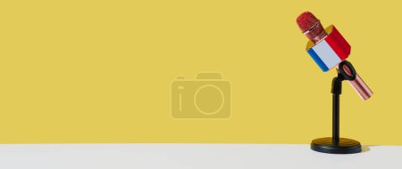 Foto de Un micrófono modelado con la bandera de Francia, sobre un soporte negro colocado sobre una superficie blanca, frente a un fondo amarillo, en un formato panorámico para utilizar como banner web - Imagen libre de derechos