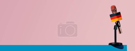Téléchargez les photos : Un microphone à motif drapeau allemand, dans un support noir, sur une surface bleue, devant un fond rose, dans un format panoramique à utiliser comme bannière web - en image libre de droit