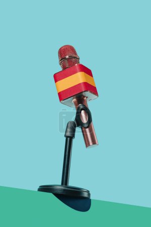 Téléchargez les photos : Gros plan d'un microphone orné du drapeau espagnol, dans un support noir placé sur une surface verte, devant un fond bleu - en image libre de droit