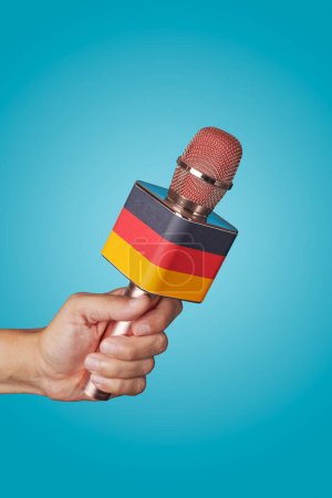 Téléchargez les photos : Gros plan de la main d'un homme tenant un microphone orné du drapeau allemand sur fond bleu - en image libre de droit