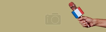 Téléchargez les photos : Un homme tient un microphone modelé avec le drapeau français, sur un fond brun avec un espace vide à gauche, dans un format panoramique à utiliser comme bannière web - en image libre de droit