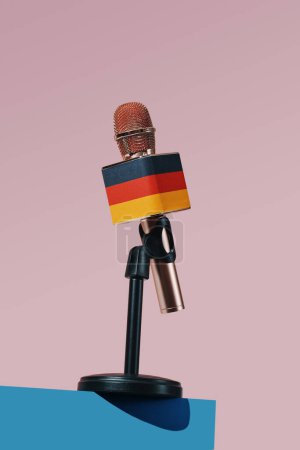 Téléchargez les photos : Gros plan d'un microphone orné du drapeau allemand, dans un support noir placé sur une surface bleue, devant un fond rose - en image libre de droit