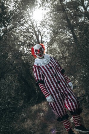 Foto de Un malvado payaso pelirrojo loco, vestido con un traje de rayas blancas y rojas con un rubor blanco, se para en el bosque mirando al observador con una sonrisa espeluznante - Imagen libre de derechos