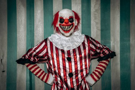 Téléchargez les photos : Un clown roux fou, portant un costume rayé blanc et rouge avec une ruche blanche, regarde l'observateur avec ses bras sur les hanches et un sourire effrayant, devant la tente du cirque - en image libre de droit