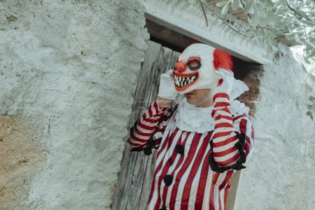 Téléchargez les photos : Un homme, vêtu d'un costume à rayures blanches et rouges à volants blancs, met ou enlève un masque de clown devant une maison rustique à l'extérieur - en image libre de droit