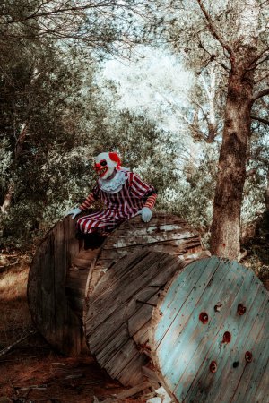 Téléchargez les photos : Un clown roux du mal fou dans les bois, portant un costume à rayures blanches et rouges avec une ruche blanche, est perché sur une bobine de câble en bois altérée au crépuscule - en image libre de droit