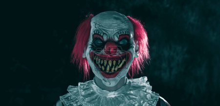 Téléchargez les photos : Un clown roux fou, portant un costume à rayures blanches et rouges avec une ruche blanche, se tient les yeux fermés et un sourire effrayant sur un fond sombre, dans un format panoramique à utiliser comme bannière web - en image libre de droit