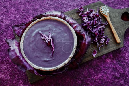 Photo for Some red cabbage soup in a rustic ceramic bowl and some chopped raw red cabbage on a wooden chopping board, placed on a purple textured surface - Royalty Free Image