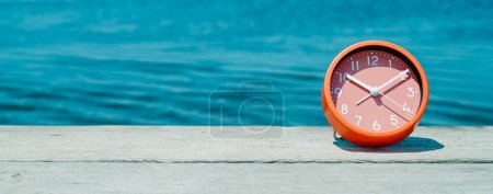 Photo for A pink clock on a weathered wooden pier, next to the water, on a sunny day, with some blank space on the left, in a panoramic format to use as web banner or header - Royalty Free Image