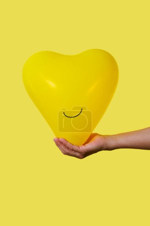 Téléchargez les photos : Homme tenant un ballon jaune souriant en forme de coeur dans sa main sur un fond jaune avec un peu d'espace vide sur le dessus et sur le fond - en image libre de droit