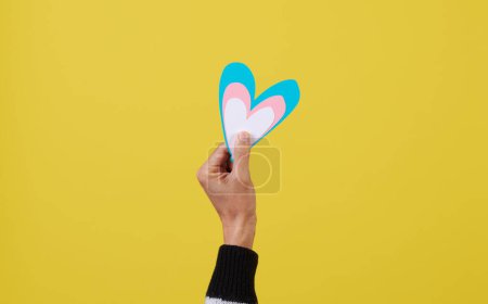 Foto de Una persona sostiene un corazón con los colores de la bandera transgénero, azul, rosa y blanco, frente a un fondo amarillo con un poco de espacio en blanco alrededor - Imagen libre de derechos