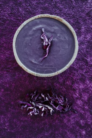 Photo for High angle view of a rustic ceramic bowl with some red cabbage soup placed on a purple textured surface next to some chopped raw red cabbage - Royalty Free Image