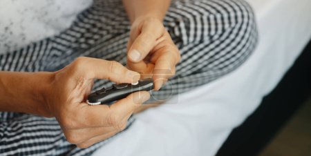 Photo for Closeup of a man sitting on his bed pricking his finger with a fingerstick to measure his blood glucose level, in a panoramic format to use as web banner - Royalty Free Image