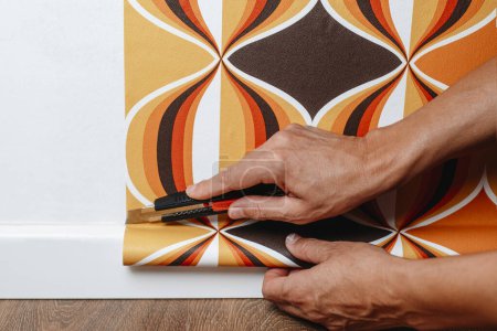 Téléchargez les photos : Gros plan d'un homme coupant les bords d'un papier peint à motifs géométriques, fraîchement fixé au mur, à côté de la plinthe blanche - en image libre de droit