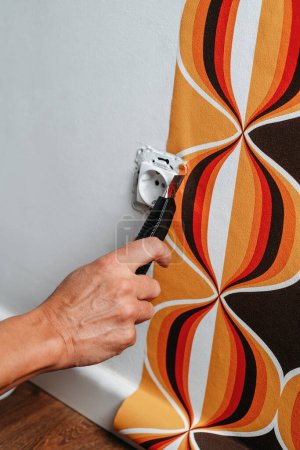 Téléchargez les photos : Un homme caucasien utilise un cutter pour couper les bords d'un papier peint à motifs géométriques autour d'une prise sur le mur - en image libre de droit