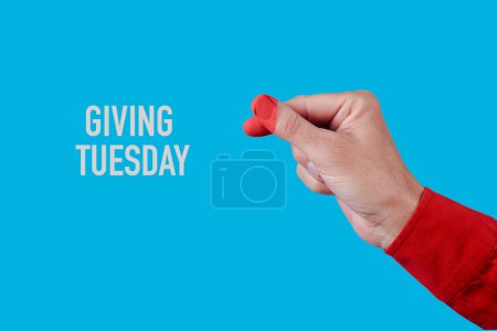 Photo for The text giving tuesday and a man doing the finger heart gesture, with the tips of his fingers painted red in the shape of a heart, on a blue background - Royalty Free Image