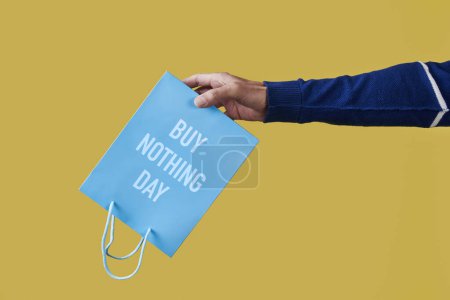 Téléchargez les photos : Gros plan d'un homme tenant un sac en papier bleu, avec le texte n'acheter rien jour écrit en elle, à l'envers sur un fond jaune - en image libre de droit