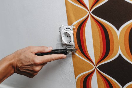 Téléchargez les photos : Gros plan d'un homme caucasien coupant les bords d'un papier peint à motifs géométriques autour d'une prise sur le mur - en image libre de droit