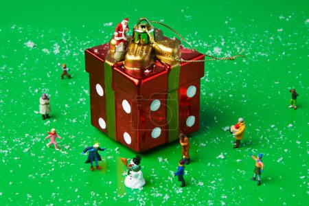 Téléchargez les photos : Un père Noël miniature se tient sur le dessus d'un ornement d'arbre de Noël en forme de cadeau, portant un sac avec des cadeaux, et un bonhomme de neige miniature et d'autres personnes miniatures jouant autour - en image libre de droit