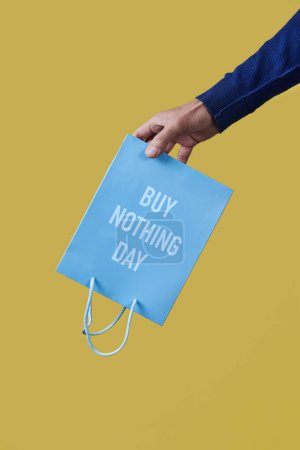 Foto de Un hombre tiene una bolsa de papel azul, con el texto comprar nada día escrito en ella, al revés en la mano, sobre un fondo amarillo - Imagen libre de derechos