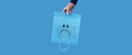Photo for A man holding a blue shopping paper bag, with a sad face painted in it, upside-down on a blue background, in a panoramic format to use as web banner - Royalty Free Image