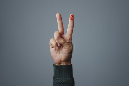 Foto de La mano de un hombre haciendo el signo V, con una cinta roja de conciencia, para la lucha contra el SIDA, pintado en su dedo índice, sobre un fondo gris con un poco de espacio en blanco alrededor - Imagen libre de derechos